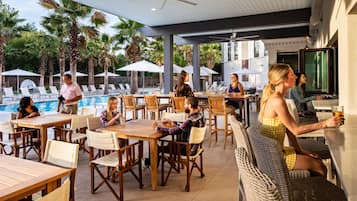 An outdoor pool area with lounge chairs and umbrellas, wooden tables, and a bar area with seating.