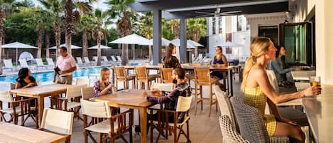 An outdoor pool area with lounge chairs and umbrellas, wooden tables, and a bar area with seating.