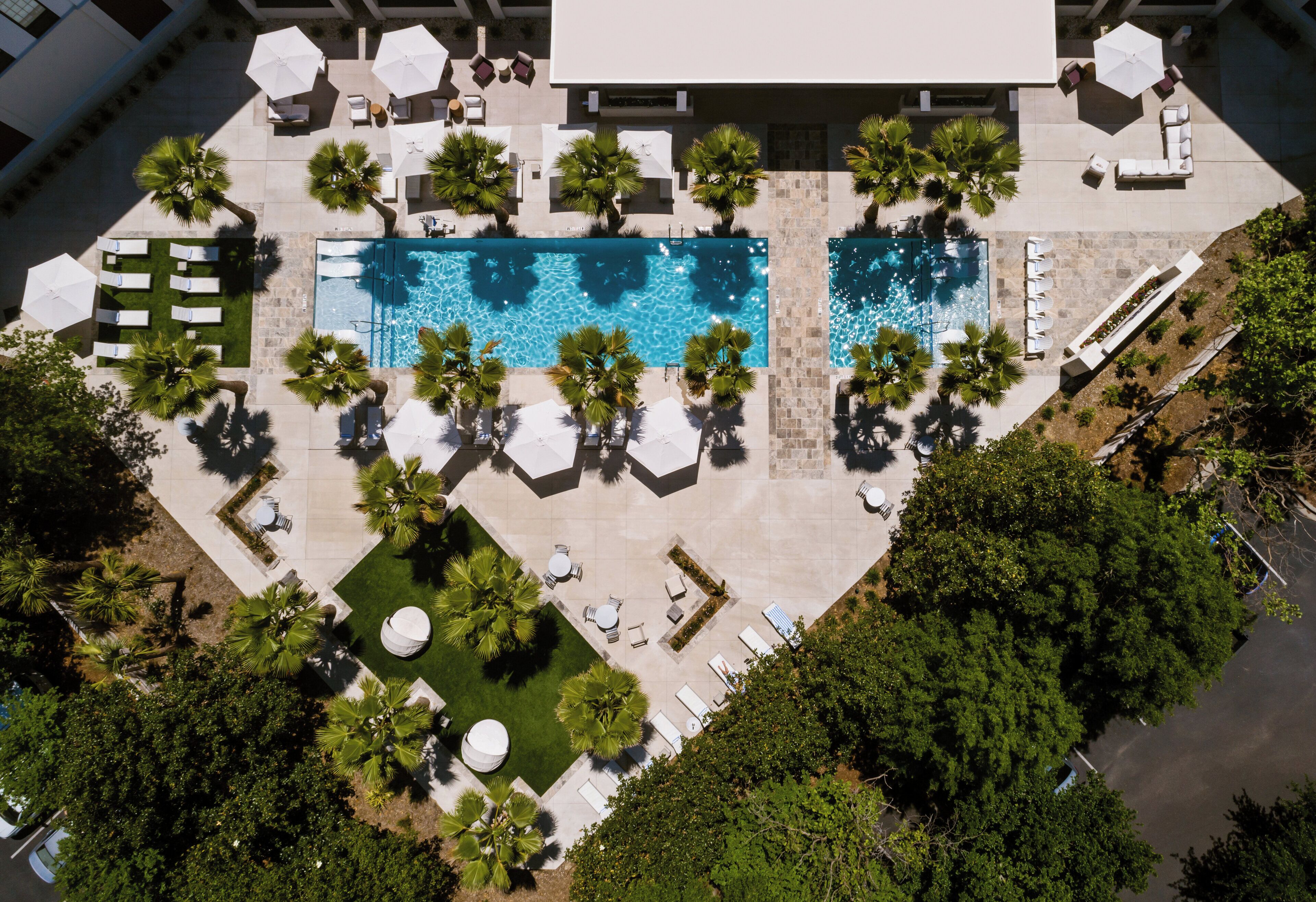 An aerial view of a resort with two swimming pools, surrounded by palm trees and sun loungers.