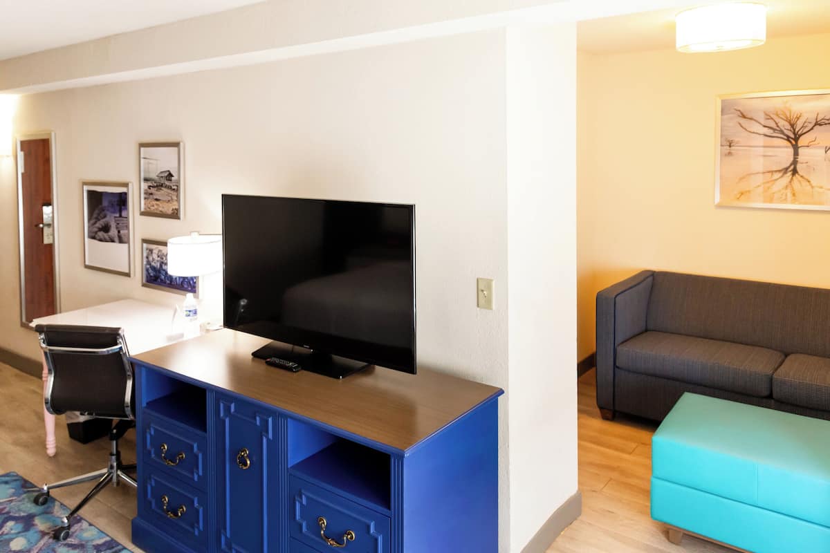 A hotel room with a flat-screen TV, a blue cabinet with drawers, a grey sofa, a teal ottoman, and a framed picture on the wall.