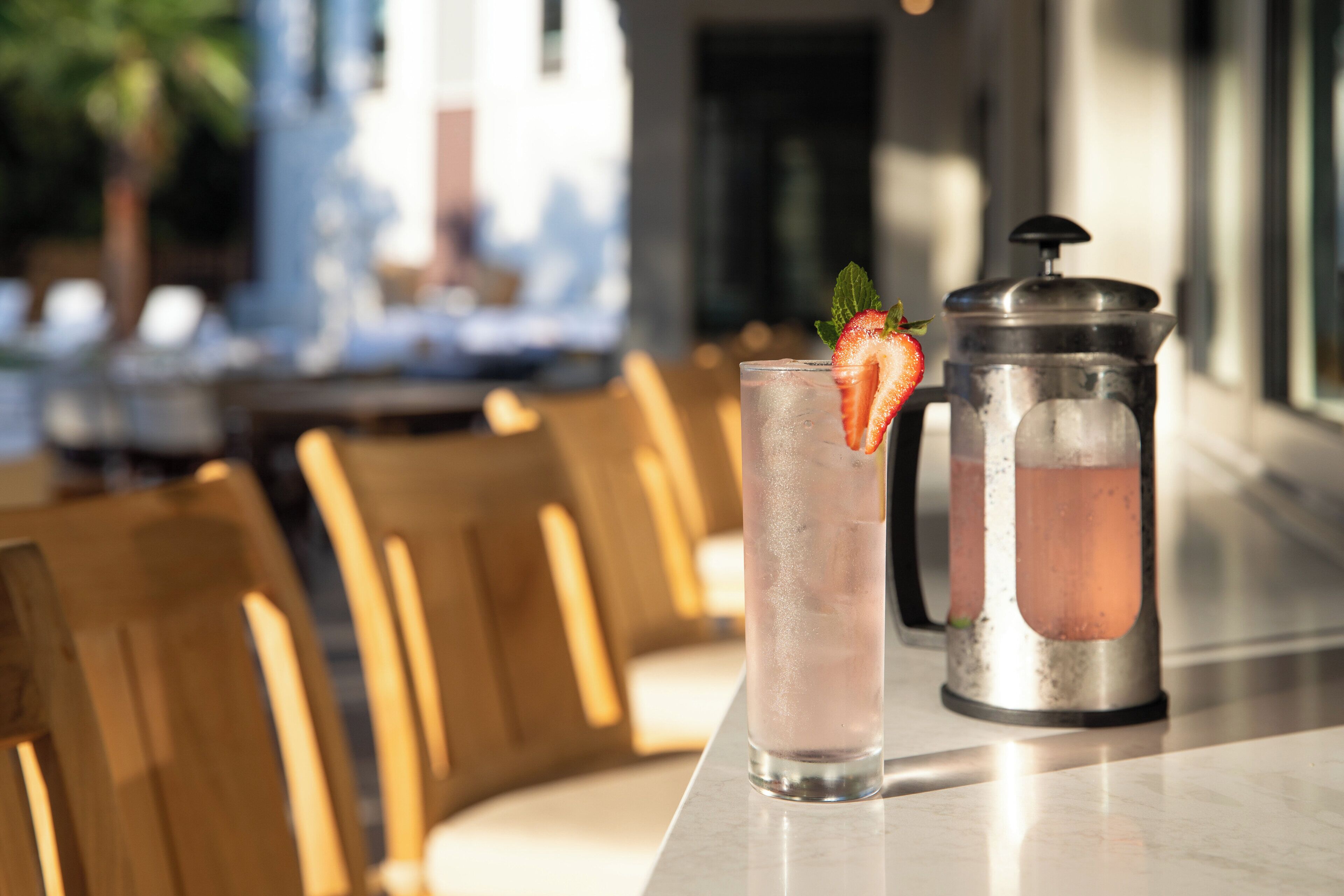 A bar setting with a tall glass of iced beverage garnished with a strawberry and mint, a metal pitcher, and wooden chairs.
