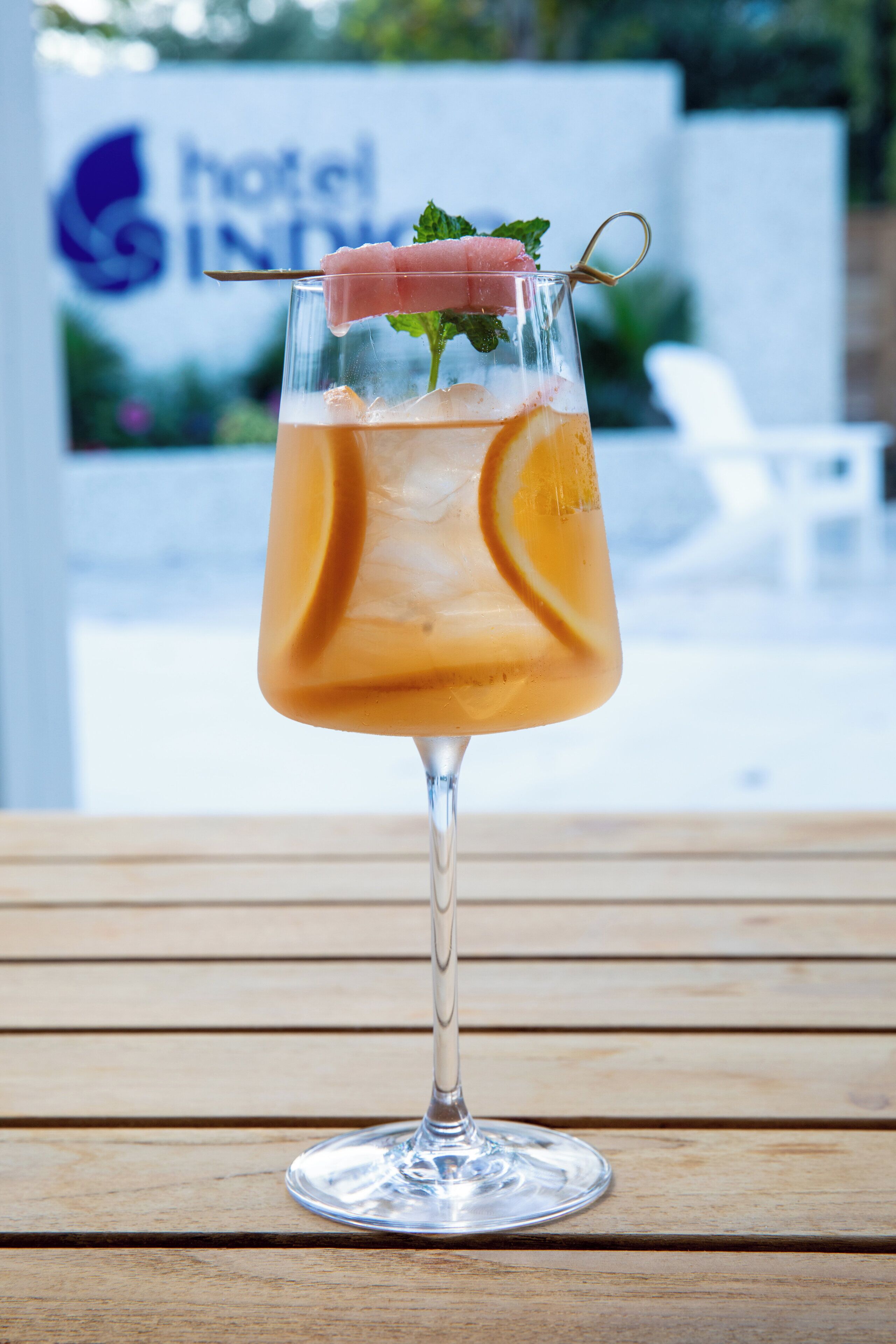 A cocktail in a stemmed glass with a garnish, ice cubes, and a slice of lemon, placed on a wooden surface.
