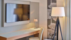 A modern hotel room with a flat-screen TV mounted on the wall, a desk with a chair, a floor lamp, and a patterned wallpaper.