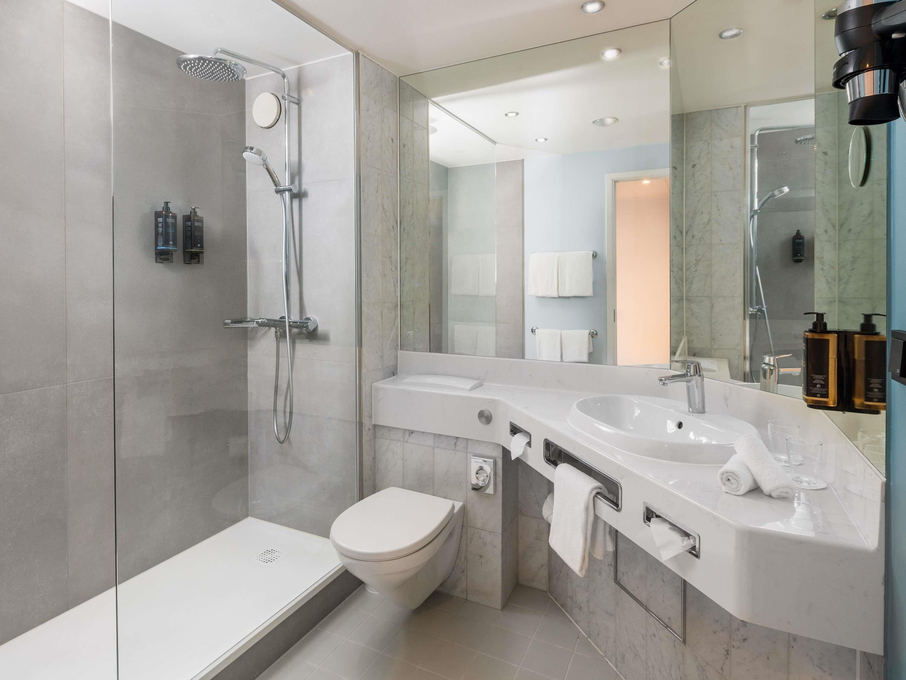 Une salle de bain moderne avec une douche à parois de verre, des toilettes blanches, un grand miroir et un meuble-lavabo doté d’un évier et d’un porte-serviettes.