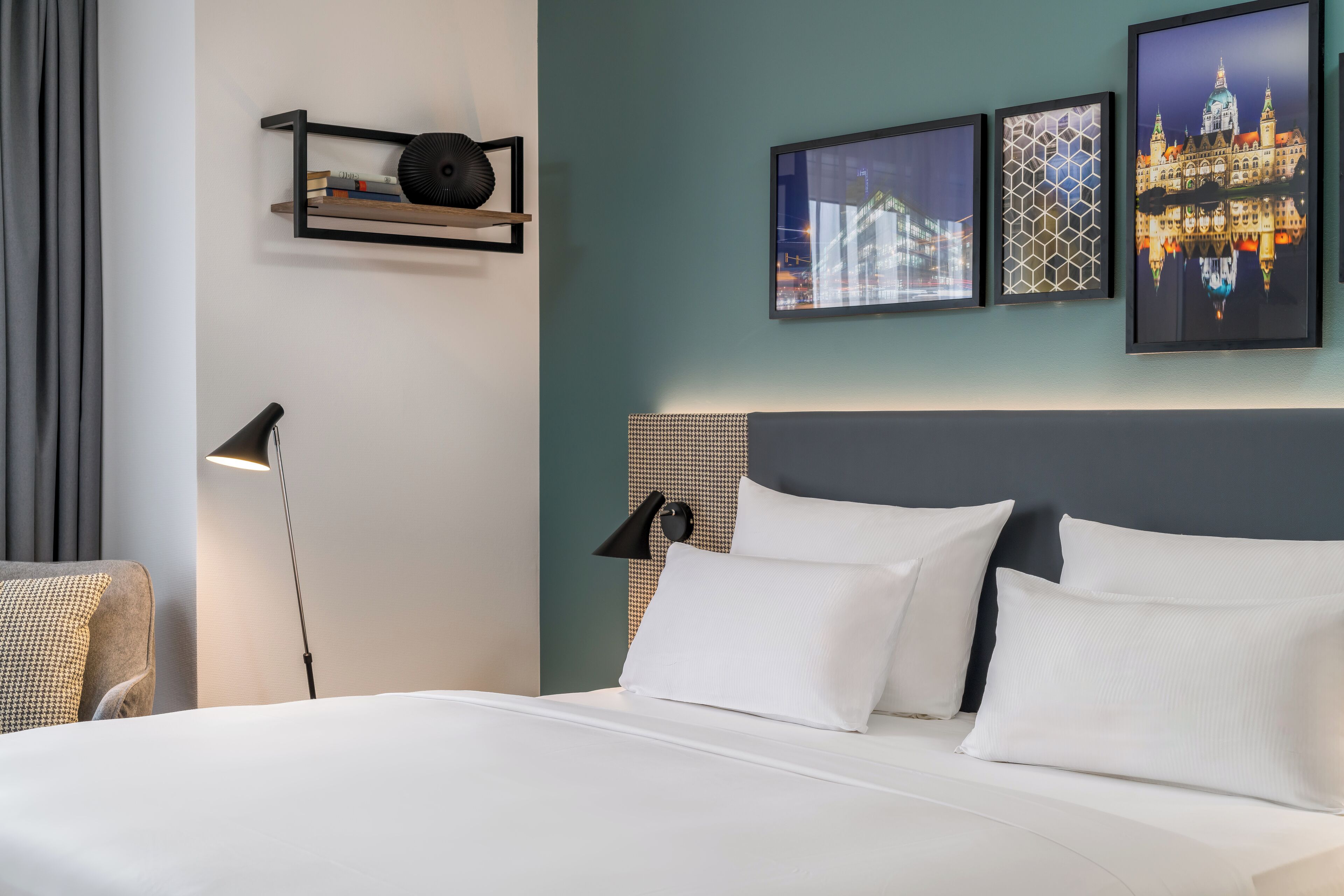 Un lit bien fait, avec du linge de lit blanc et une tête de lit, une lampe de chevet et une étagère avec des livres et un objet décoratif.