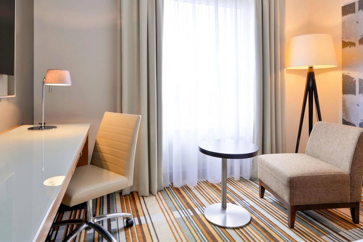 Une chambre d’hôtel équipée d’un bureau, de deux chaises, d’une petite table ronde, d’un lampadaire et d’une grande fenêtre avec des rideaux.