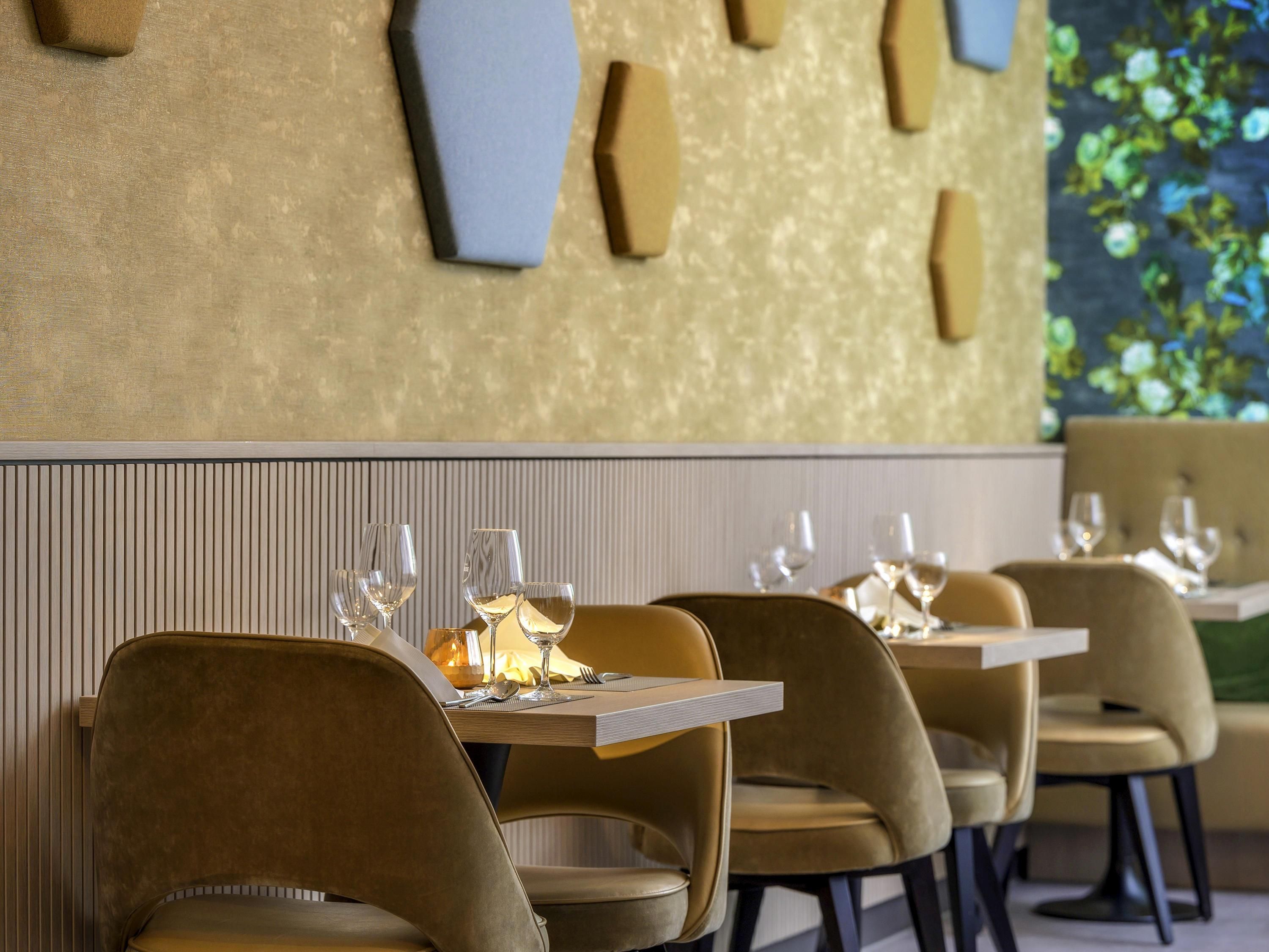 A restaurant interior with a modern design, featuring a long table with multiple chairs, a decorative wall, and a view of greenery outside.