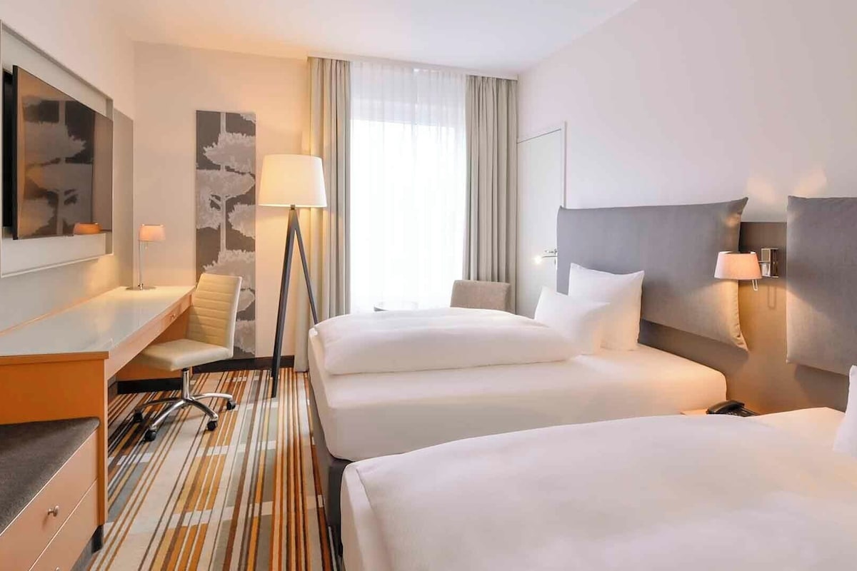 A hotel room with two beds, a desk, a chair, a lamp, and a large window with curtains.