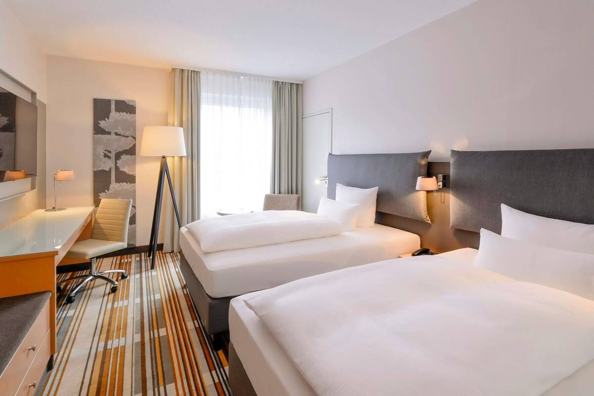 Habitación de hotel con dos camas, un escritorio, una silla, una lámpara y un ventanal grande con cortinas.