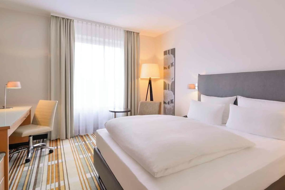 A hotel room with a large bed, a desk with a chair, a lamp, and a view of the outdoors through the window.