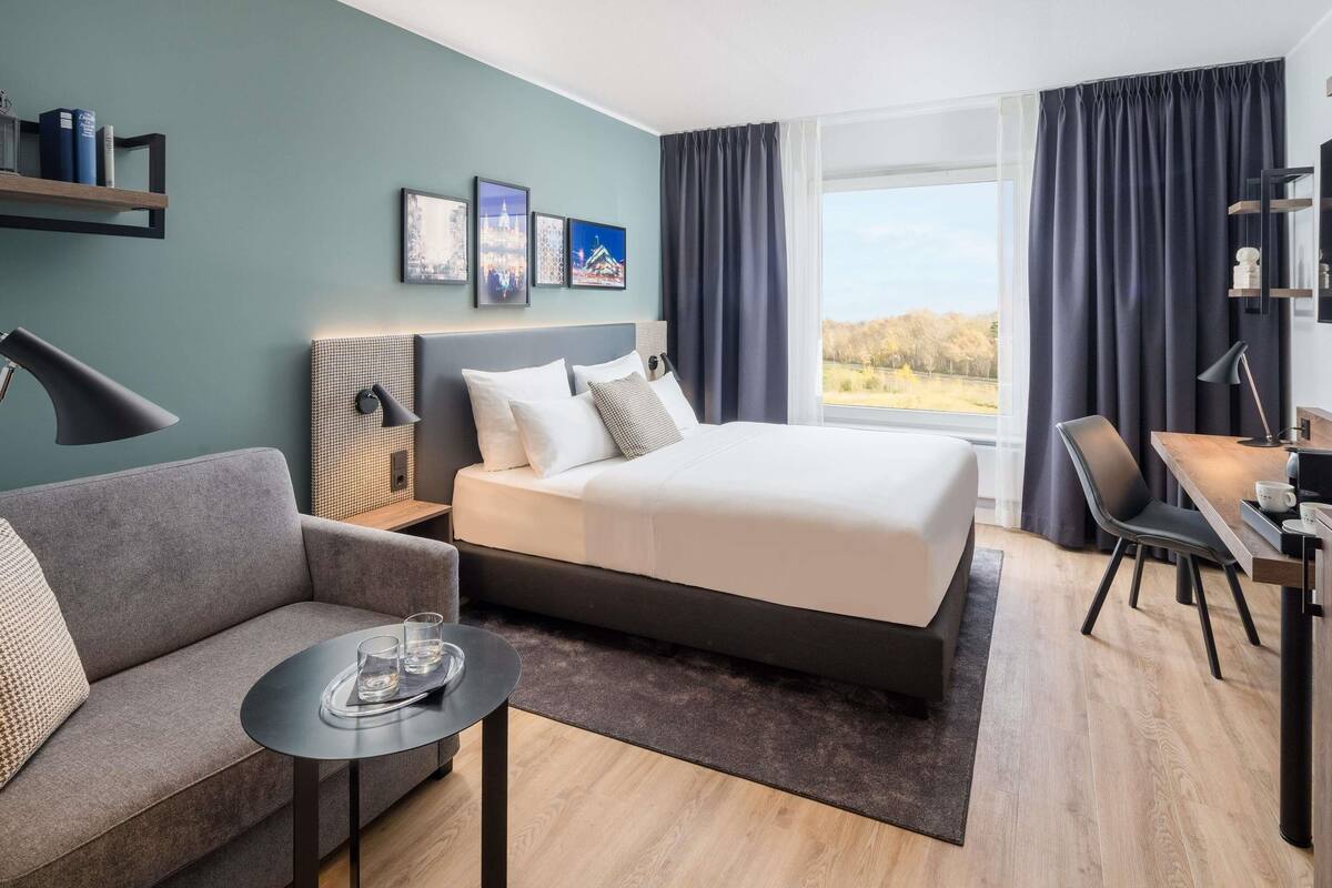 Una habitación de hotel moderna con una cama grande, un sofá, una mesita y un escritorio. Hay cuadros en la pared, una ventana con cortinas y vistas al exterior.