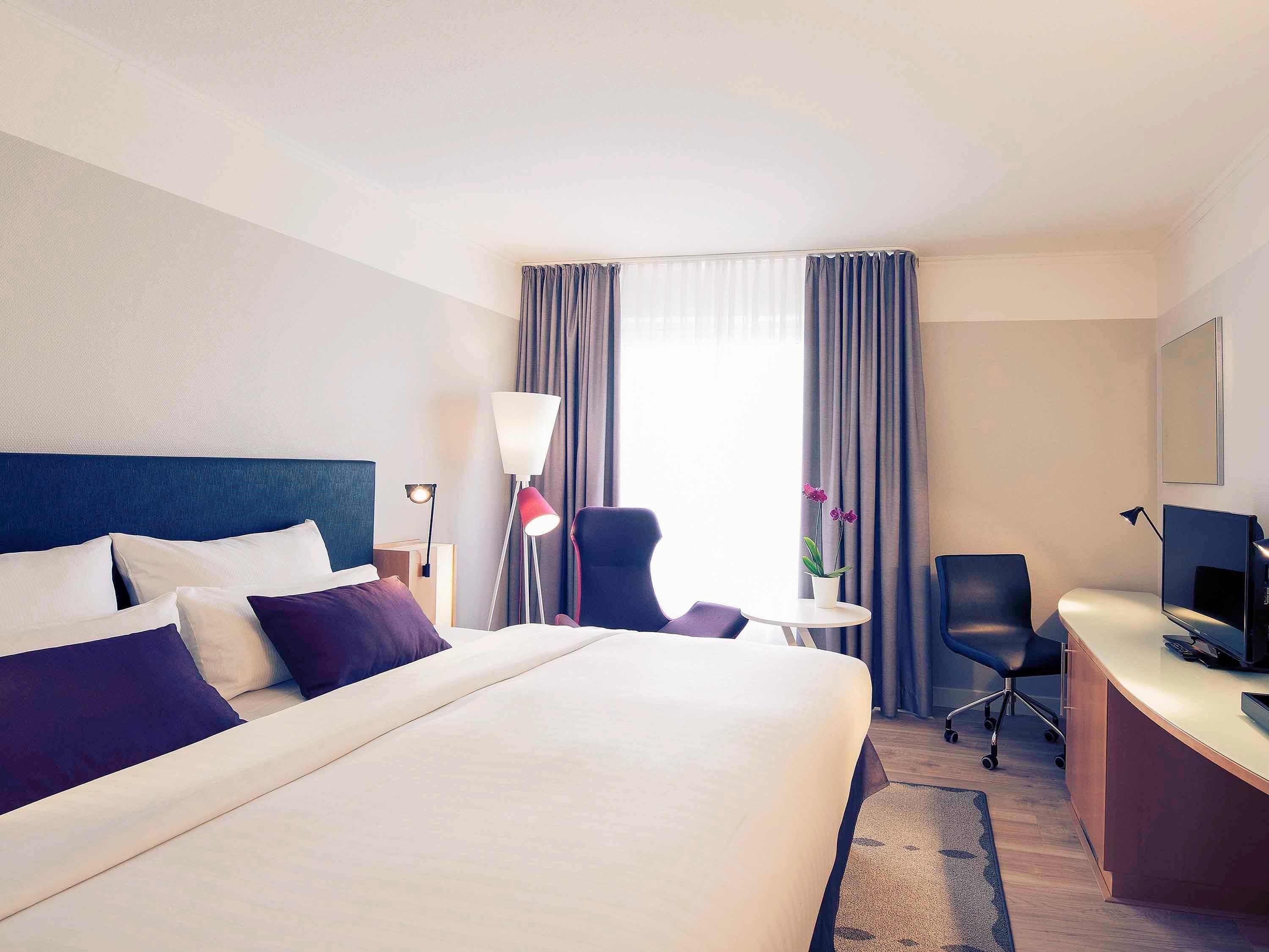A hotel room with a large bed, a desk with a chair, a lamp, and a TV.