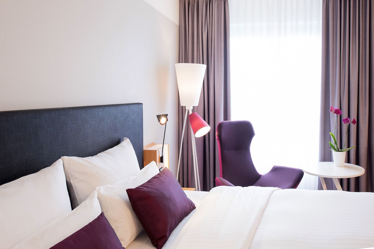 A hotel room with a bed, a chair, a small table, and a lamp.
