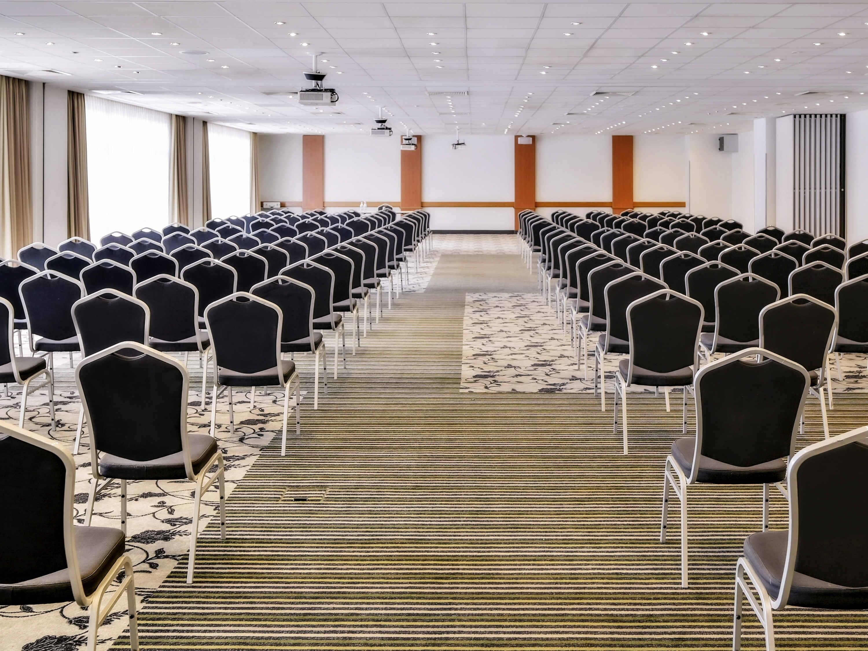 Un salón de conferencias con filas de sillas, piso alfombrado y un proyector en el techo.
