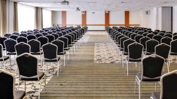 Un salón de conferencias con filas de sillas, piso alfombrado y un proyector en el techo.