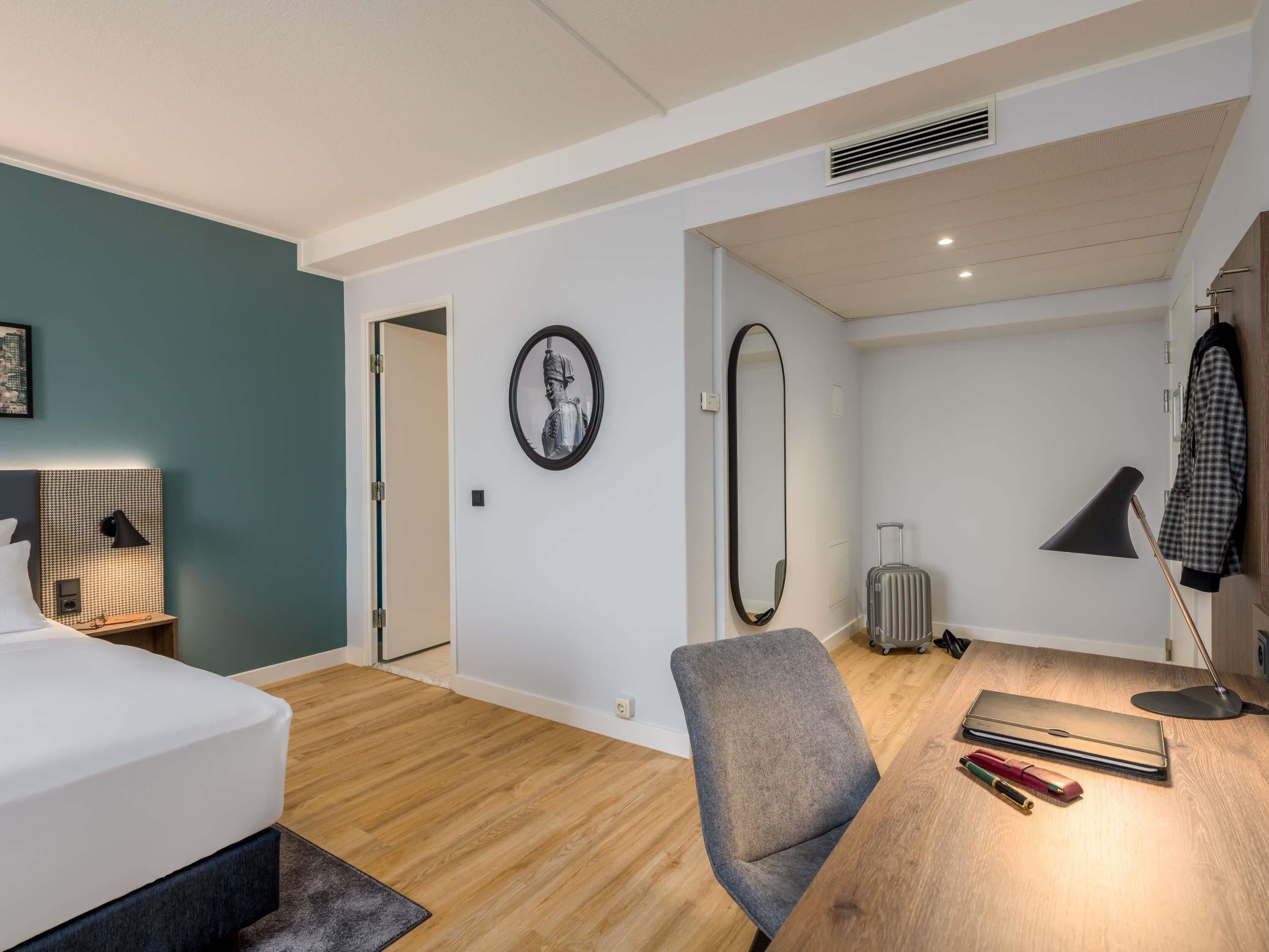 A modern hotel room with a bed, a desk, a chair, a mirror, and a suitcase.