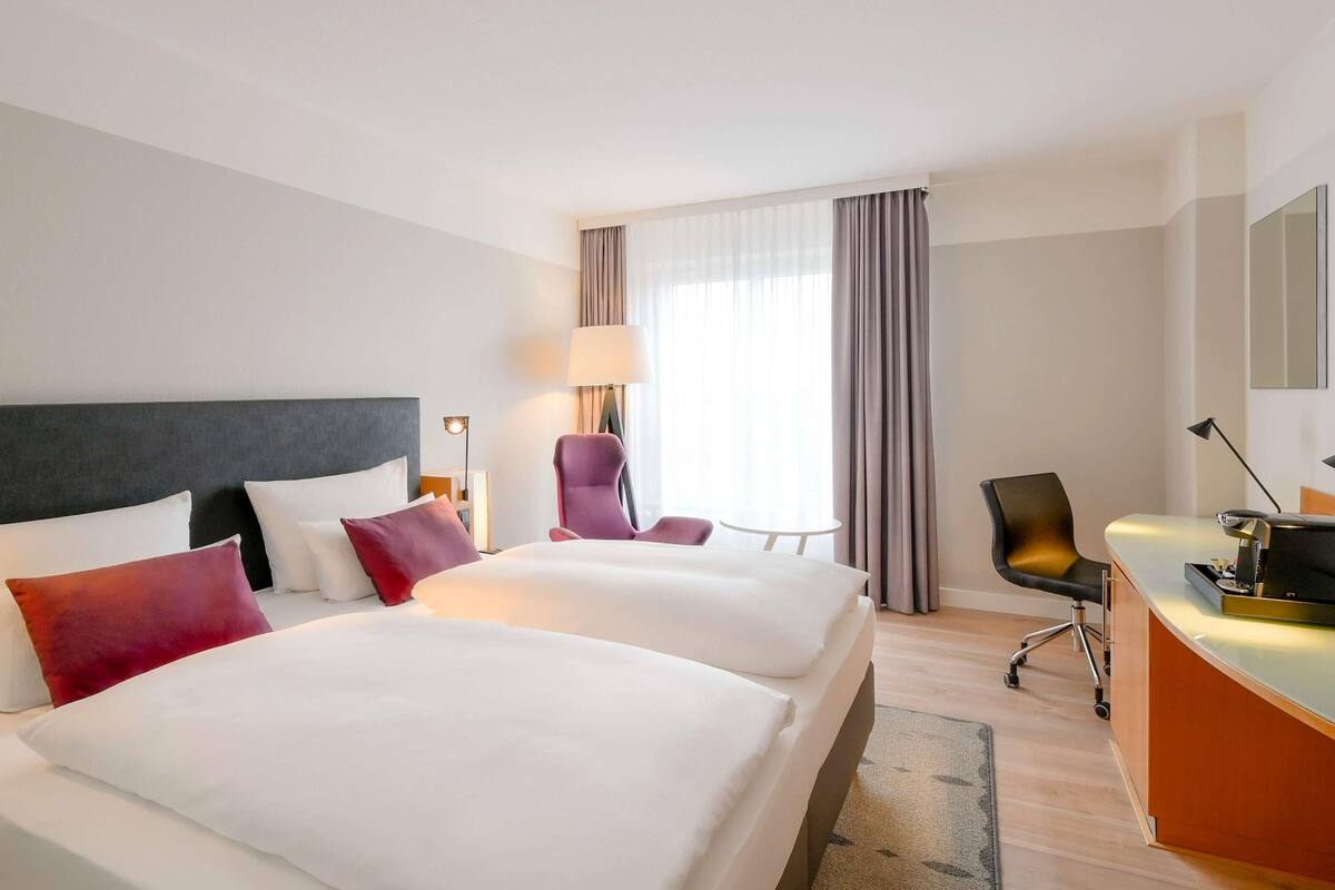 Habitación de hotel con dos camas, un escritorio, una silla, una lámpara y un ventanal grande con cortinas.