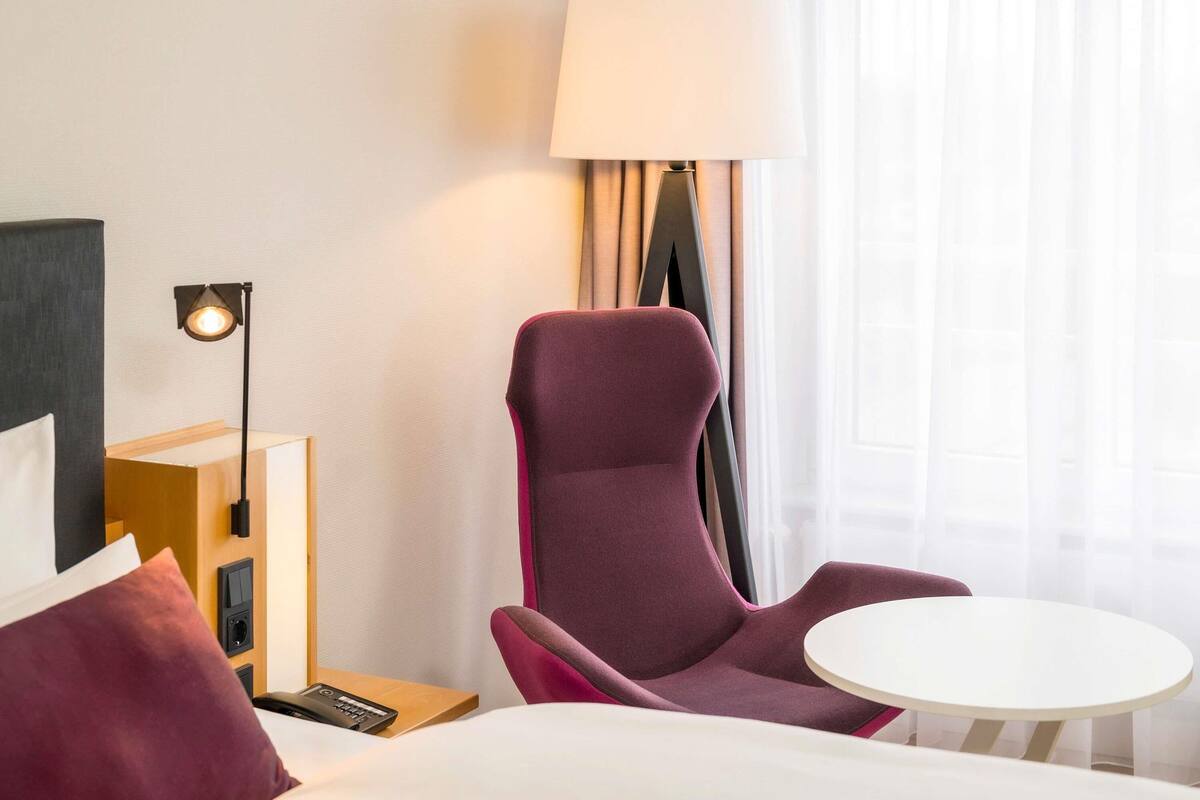 Une chambre d’hôtel avec un lit, un fauteuil violet, une petite table ronde, une lampe et une table de chevet avec un téléphone et une télécommande.