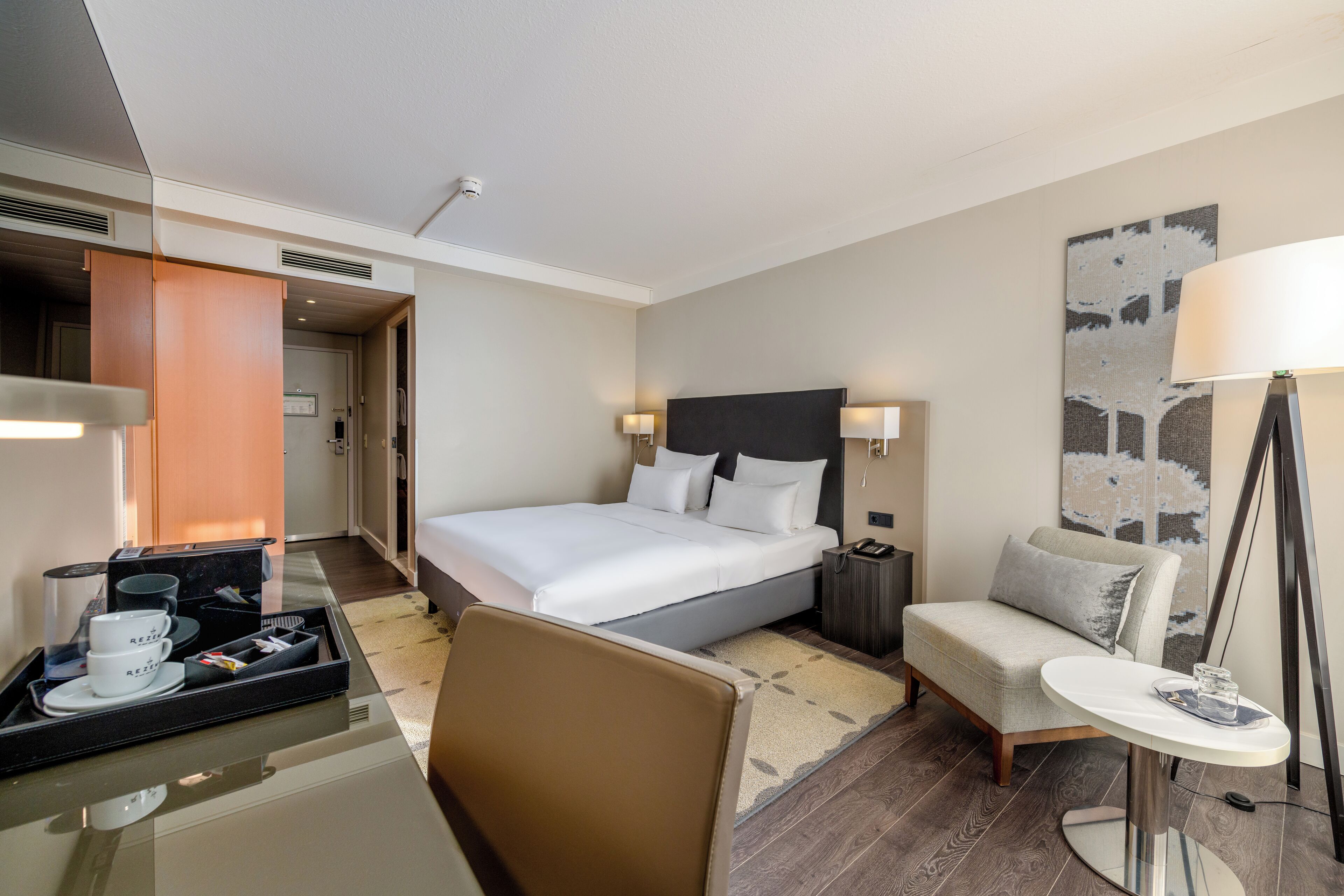 A hotel room with a bed, bedside table, chair, desk, and a large window.