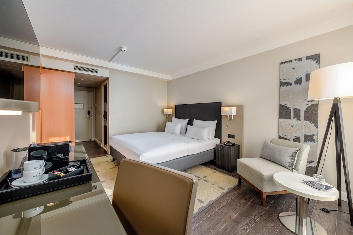A hotel room with a bed, bedside table, chair, desk, and a large window.