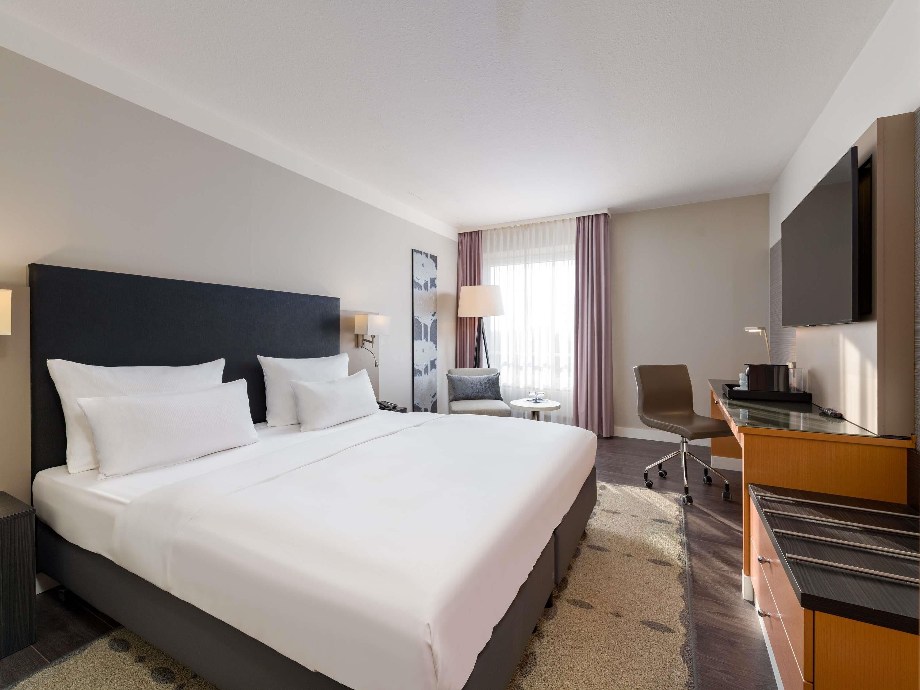 A hotel room with a large bed, a desk, a chair, a TV, and a window with curtains.