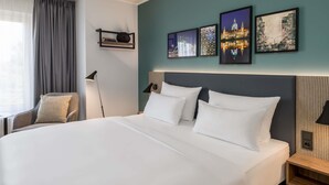 A hotel room with a large bed, a desk, a chair, and a window with curtains. There are four framed pictures on the wall above the bed.