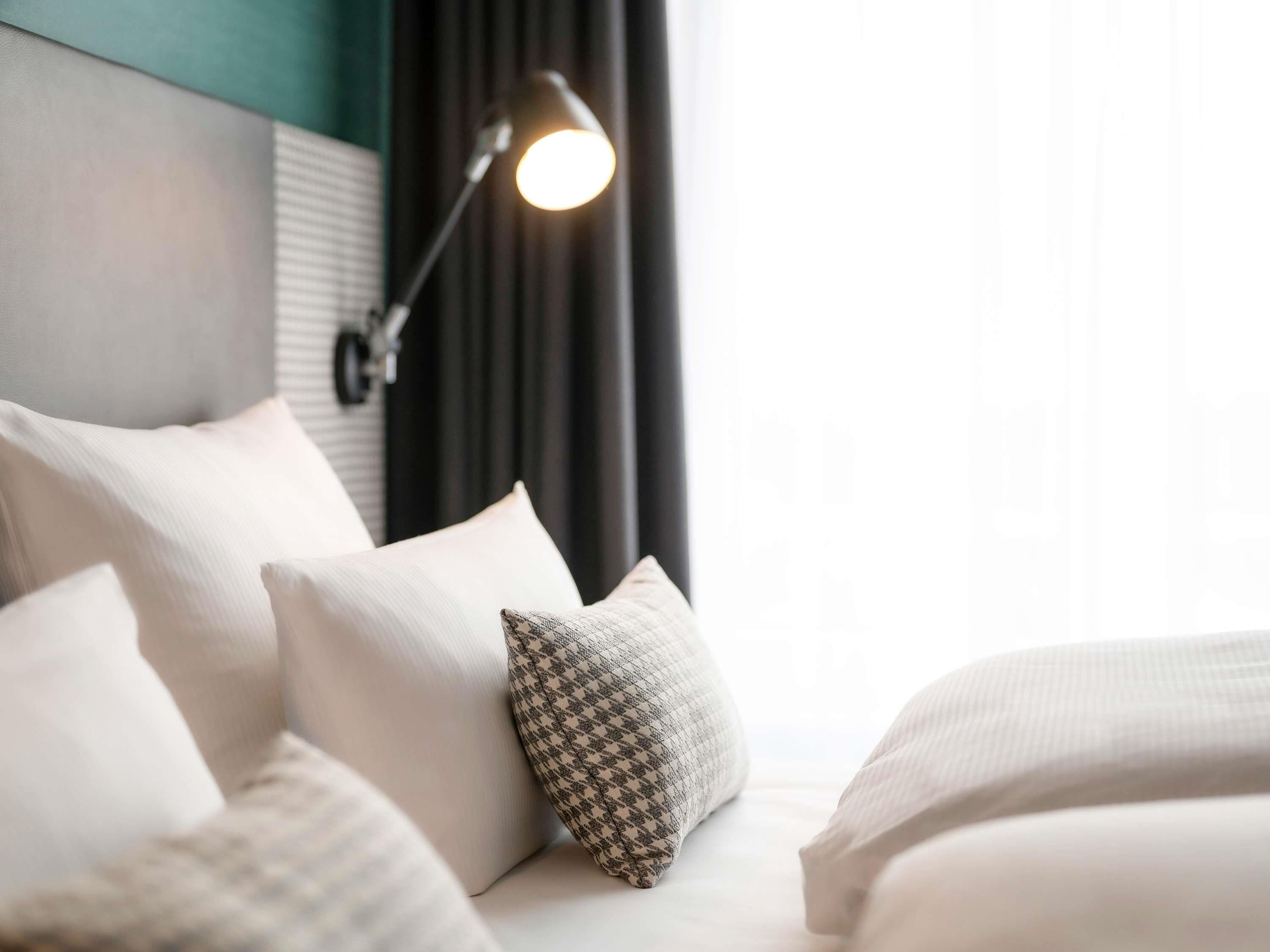 A neatly made bed with white bedding and a patterned pillow, a bedside lamp, and a window with curtains.
