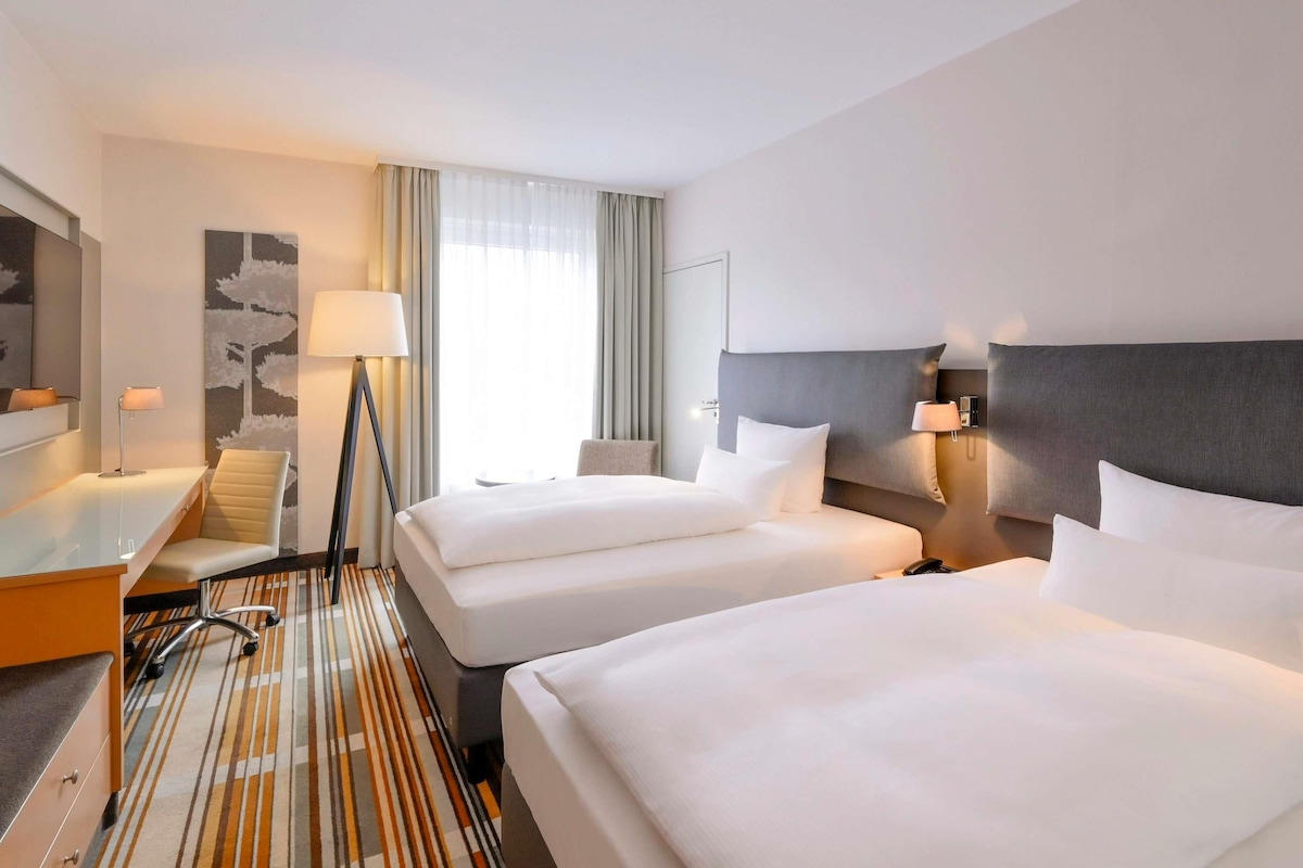 Habitación de hotel con dos camas, un escritorio, una silla, una lámpara y un ventanal grande con cortinas.