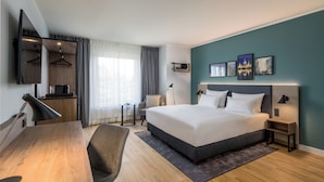 A modern hotel room with a large bed, a desk, a chair, a TV, and artwork on the wall.