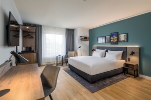 Una habitación de hotel moderna con una cama grande, un escritorio, una silla, un televisor y obras de arte en la pared.