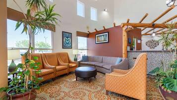 A spacious lobby with a patterned carpet, multiple seating areas including a sofa and armchairs, a coffee table, and a large potted plant.