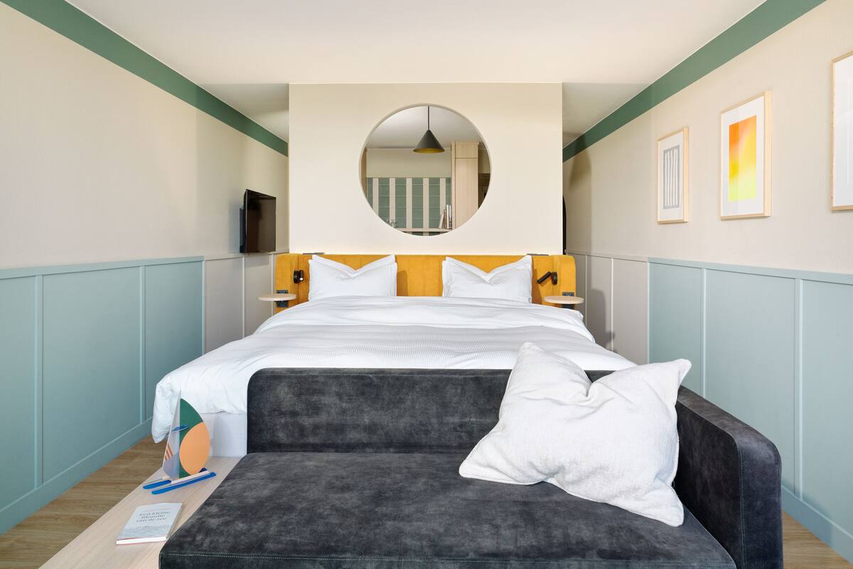 A hotel room with a bed, a sofa, a round mirror, and framed pictures on the wall.