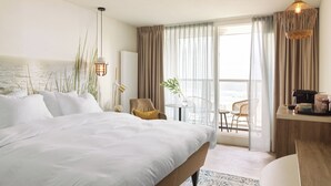 A hotel room with a large bed, a desk with a computer, a chair, and a view of the sea through the window. - Carlton Beach (The Hague)