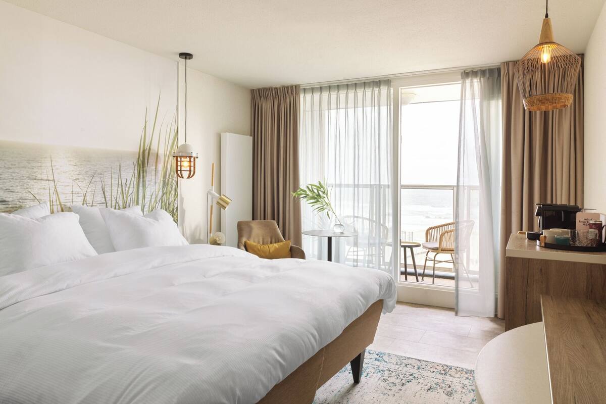 A hotel room with a large bed, a desk with a computer, a chair, and a view of the sea through the window.