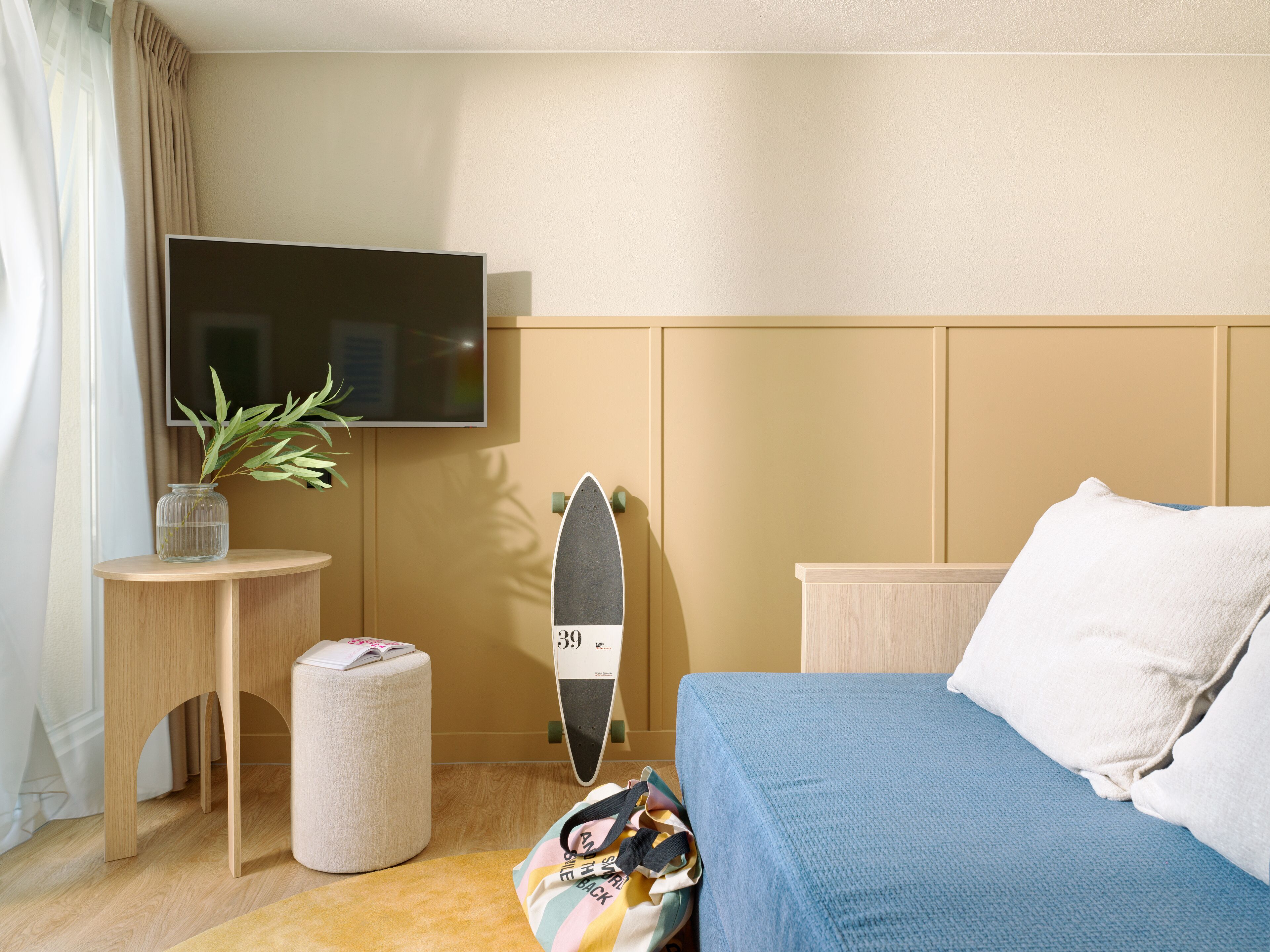 Habitación de hotel con cama, televisor, una mesita con una planta, un monopatín y un bolso colorido.