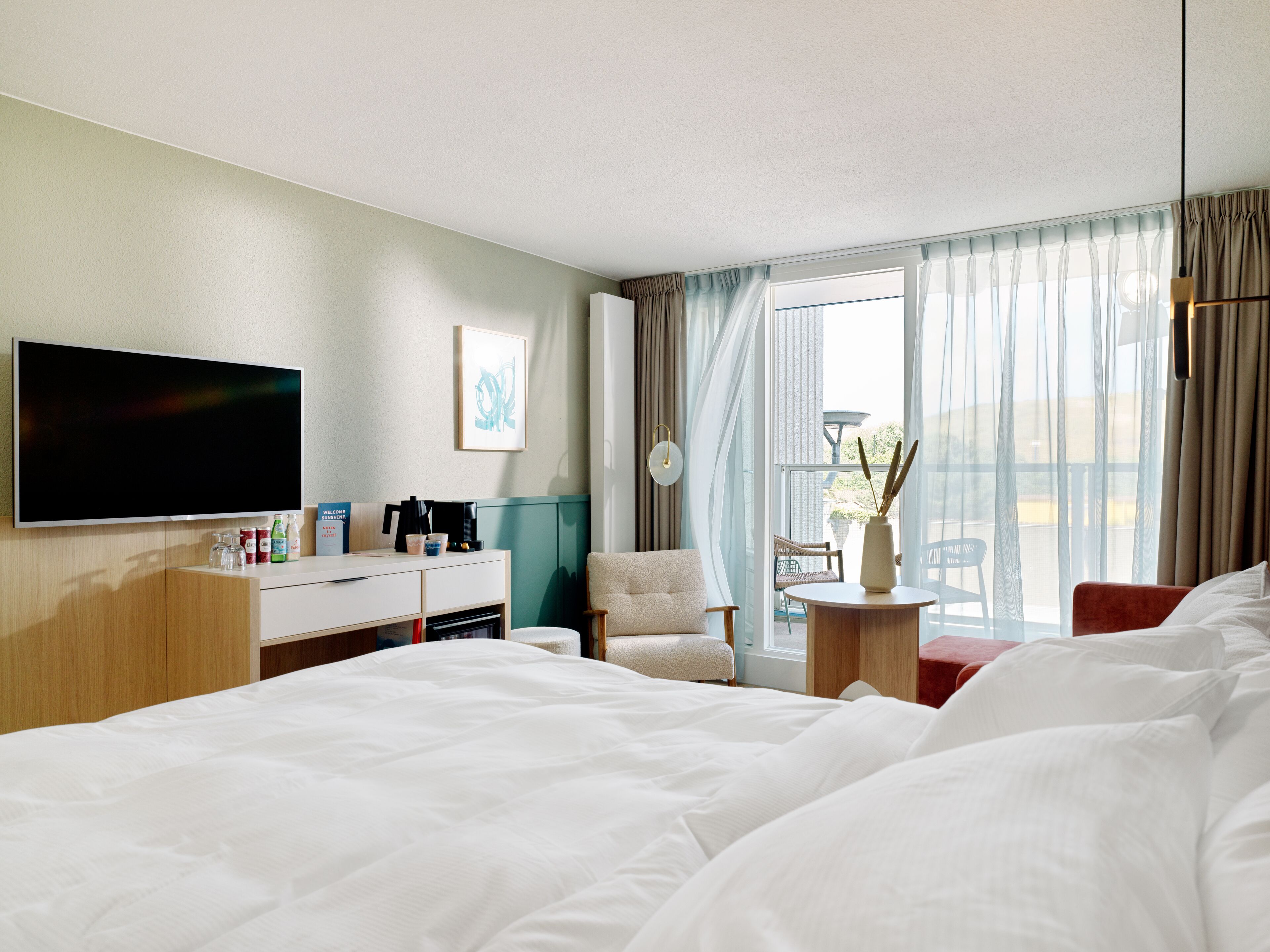 A hotel room with a large bed, a flat-screen TV, a desk with a coffee maker, and a view of the outdoors through the window.