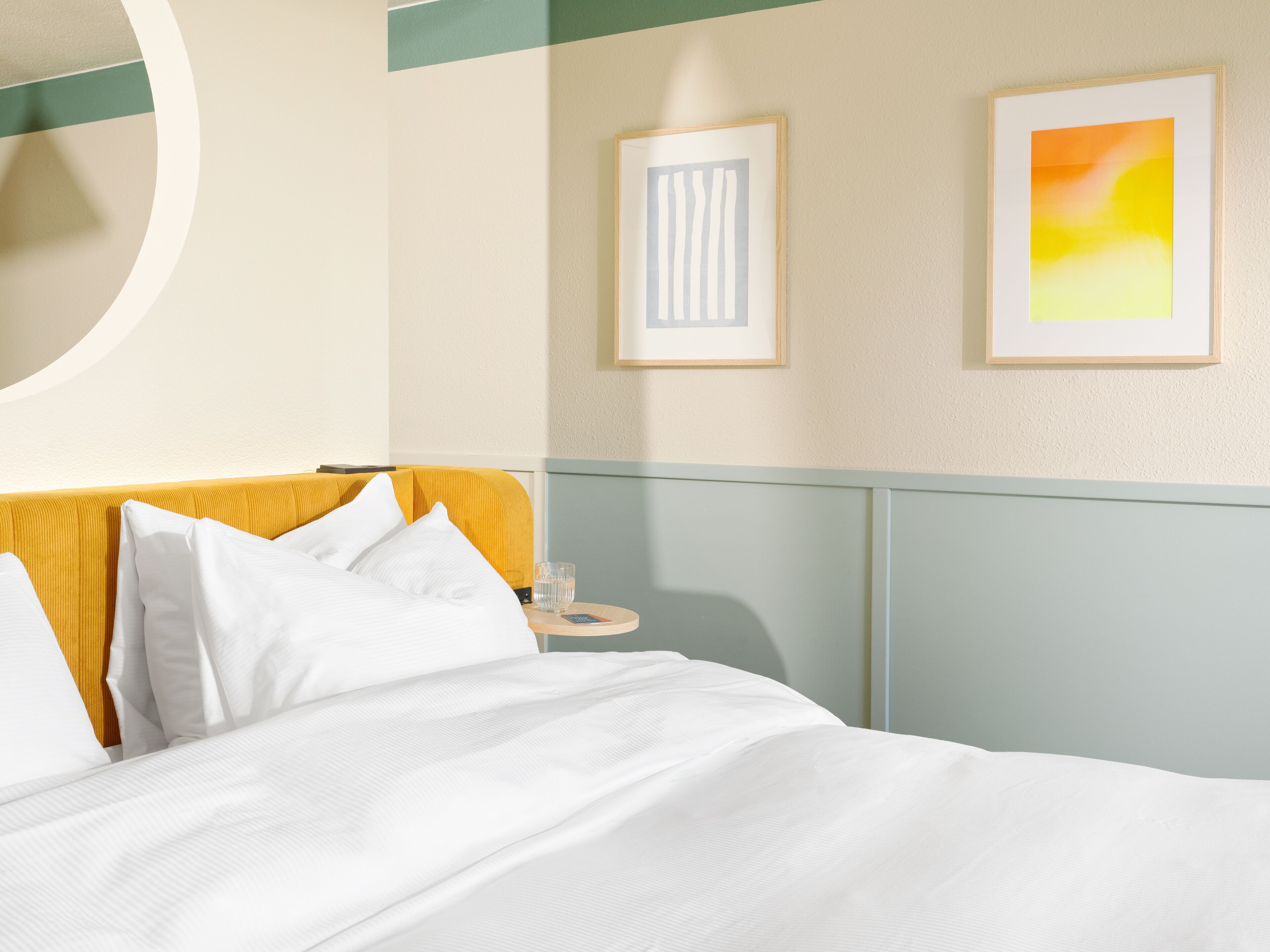 A hotel room with a bed, a bedside table, and two framed artworks on the wall.