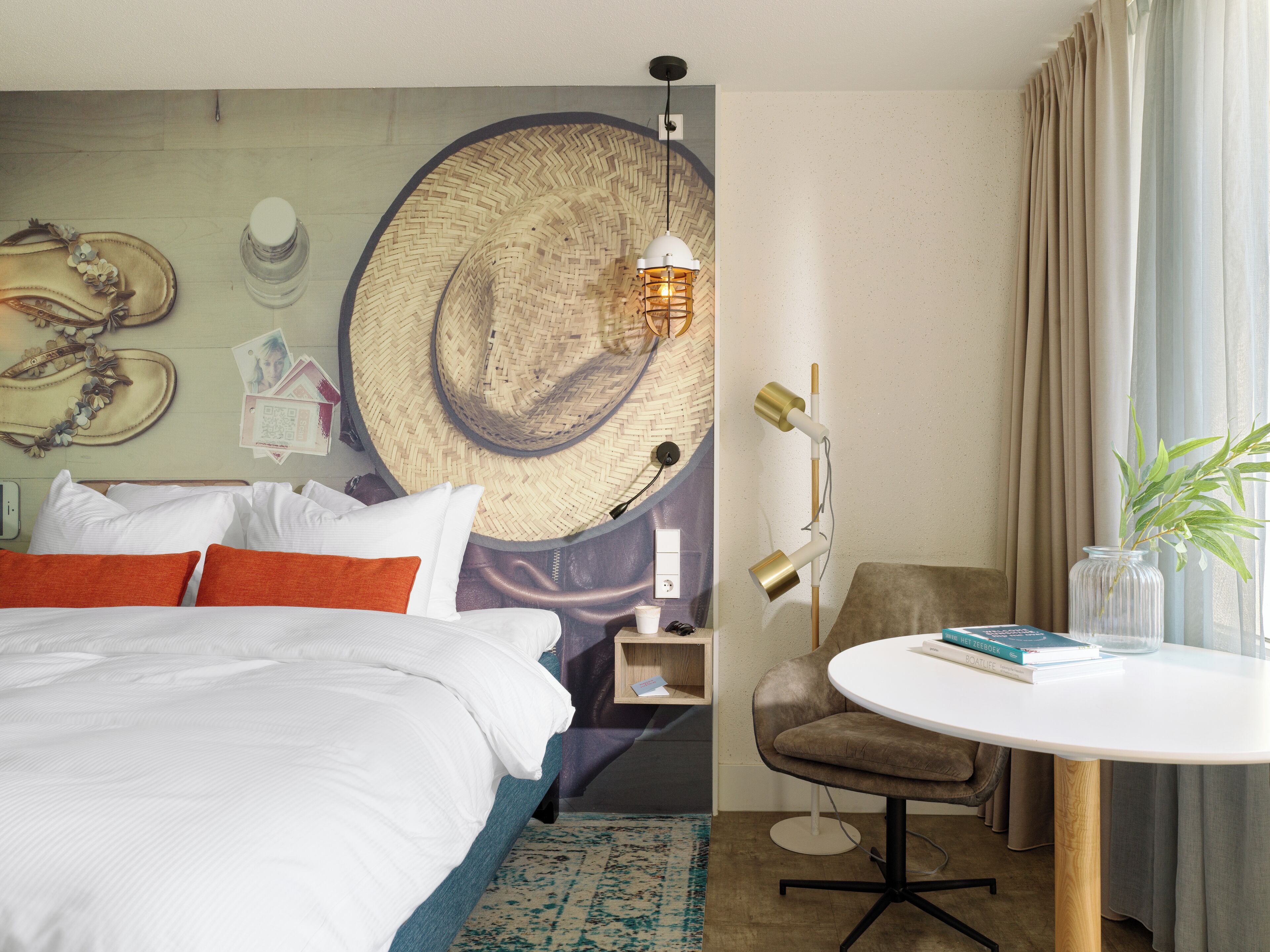 Habitación de hotel con cama, una mesa redonda, una silla, una lámpara y una pared decorada con un sombrero tejido y un reloj de pared.
