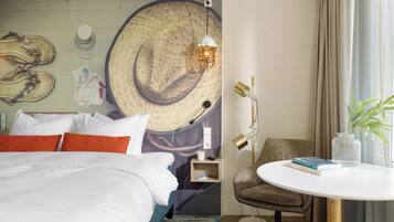 Habitación de hotel con cama, una mesa redonda, una silla, una lámpara y una pared decorada con un sombrero tejido y un reloj de pared.