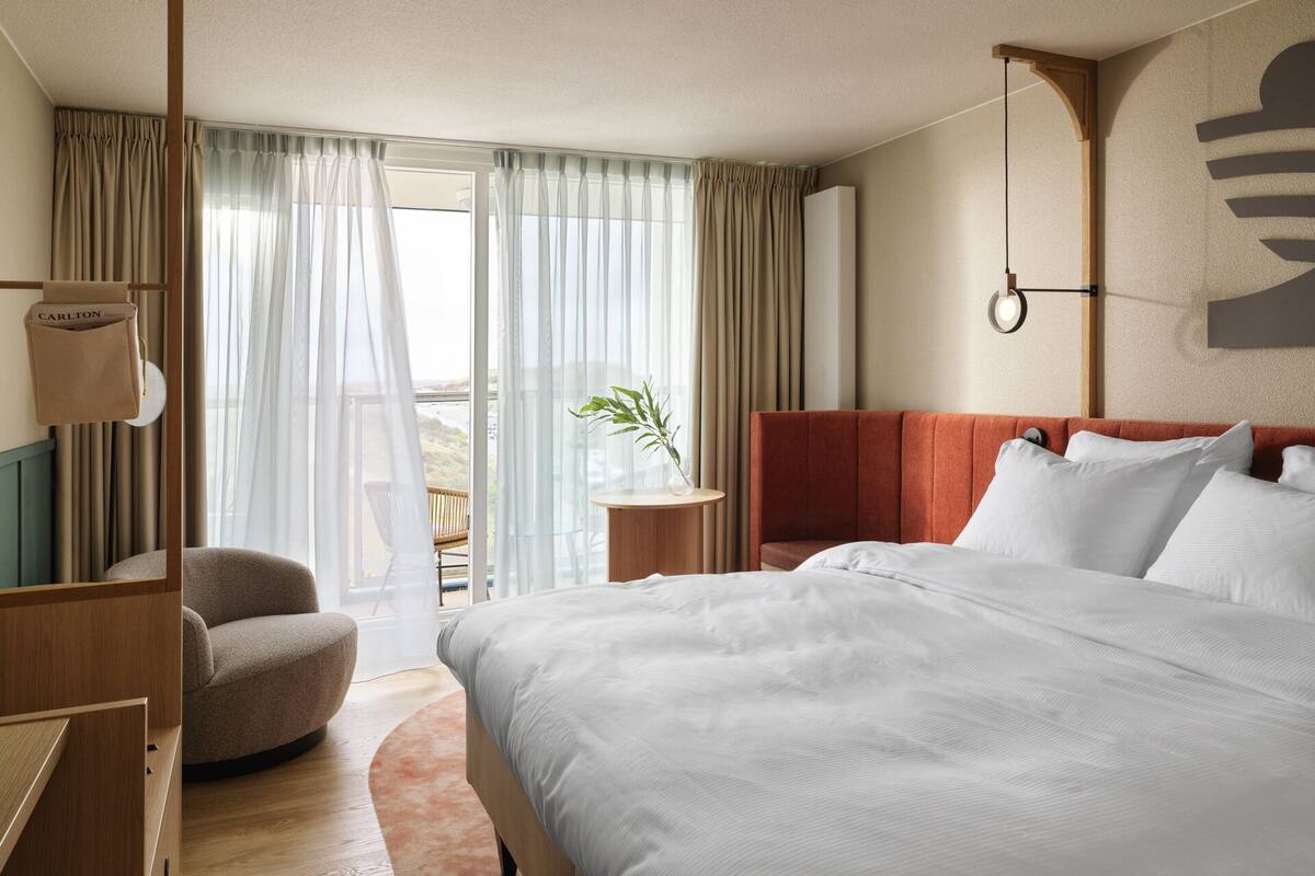 A hotel room with a large bed, a wooden nightstand, a chair, a small table with a plant, and a view of the outdoors through sheer curtains.