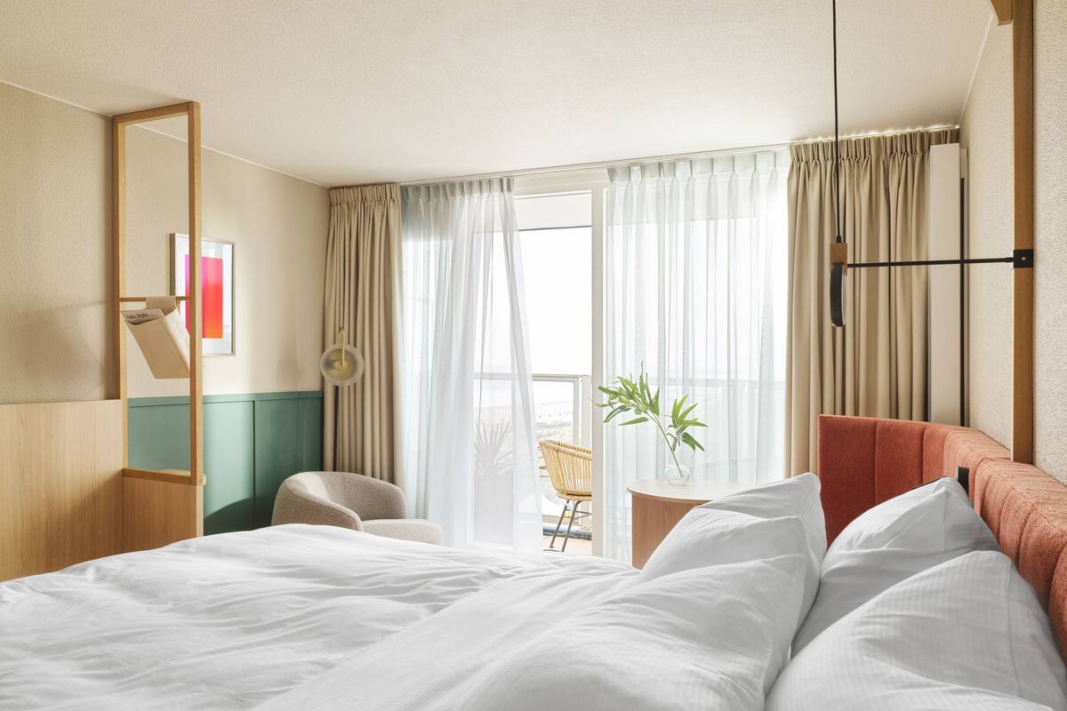 A hotel room with a bed, a chair, a small table with a plant, and a window with curtains.
