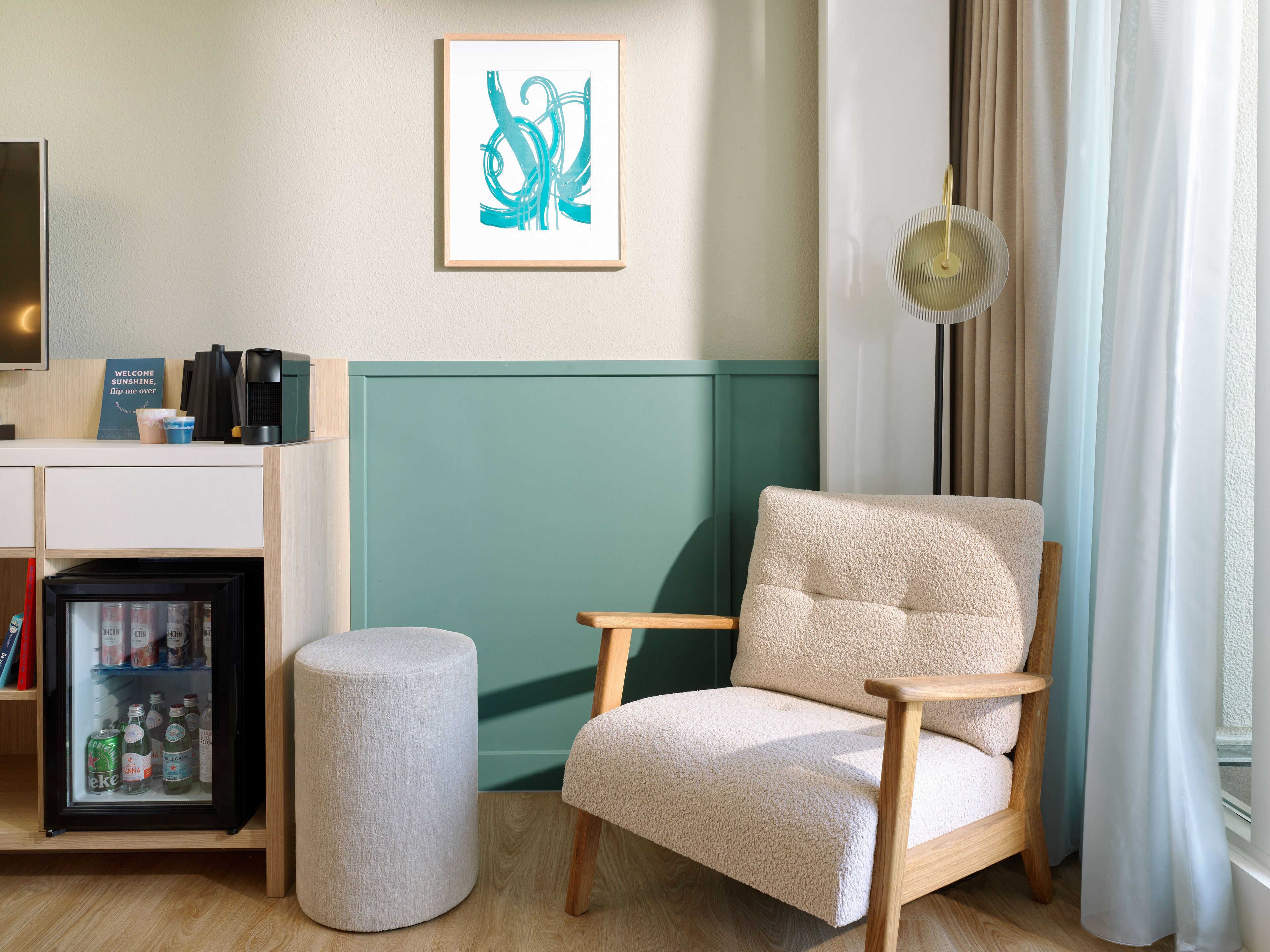 A modern room with a teal accent wall, a plush armchair, a small round ottoman, a minibar, and a framed abstract artwork.