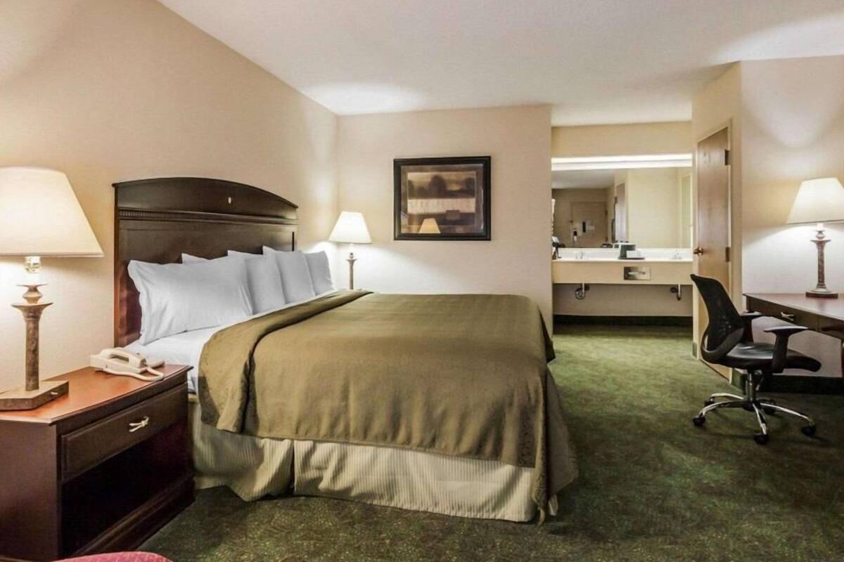 A hotel room with a bed, bedside lamps, a desk with a chair, a mirror, and a framed picture on the wall.