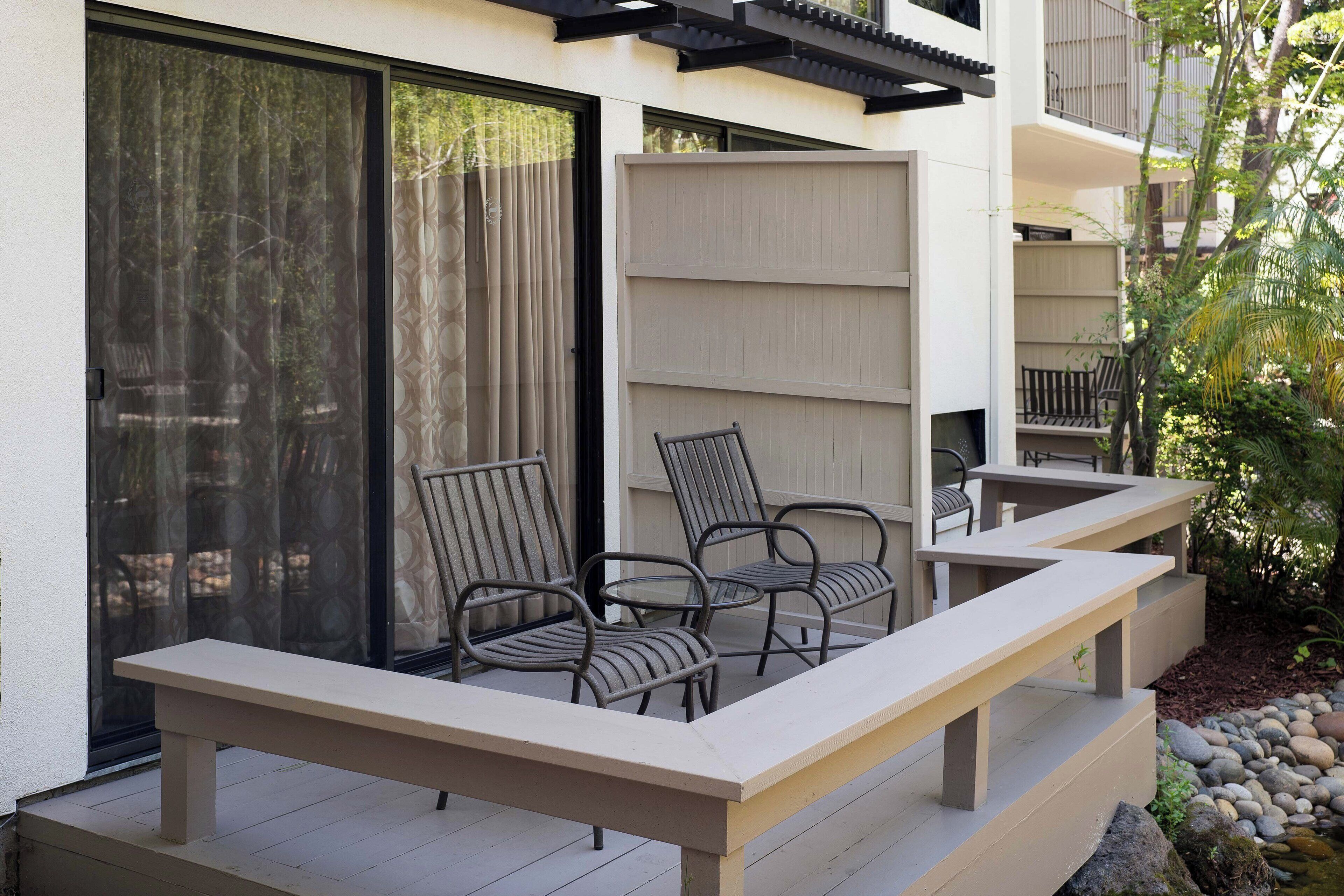 A balcony with a bench and two chairs, a sliding glass door, and a small garden area with rocks and plants.