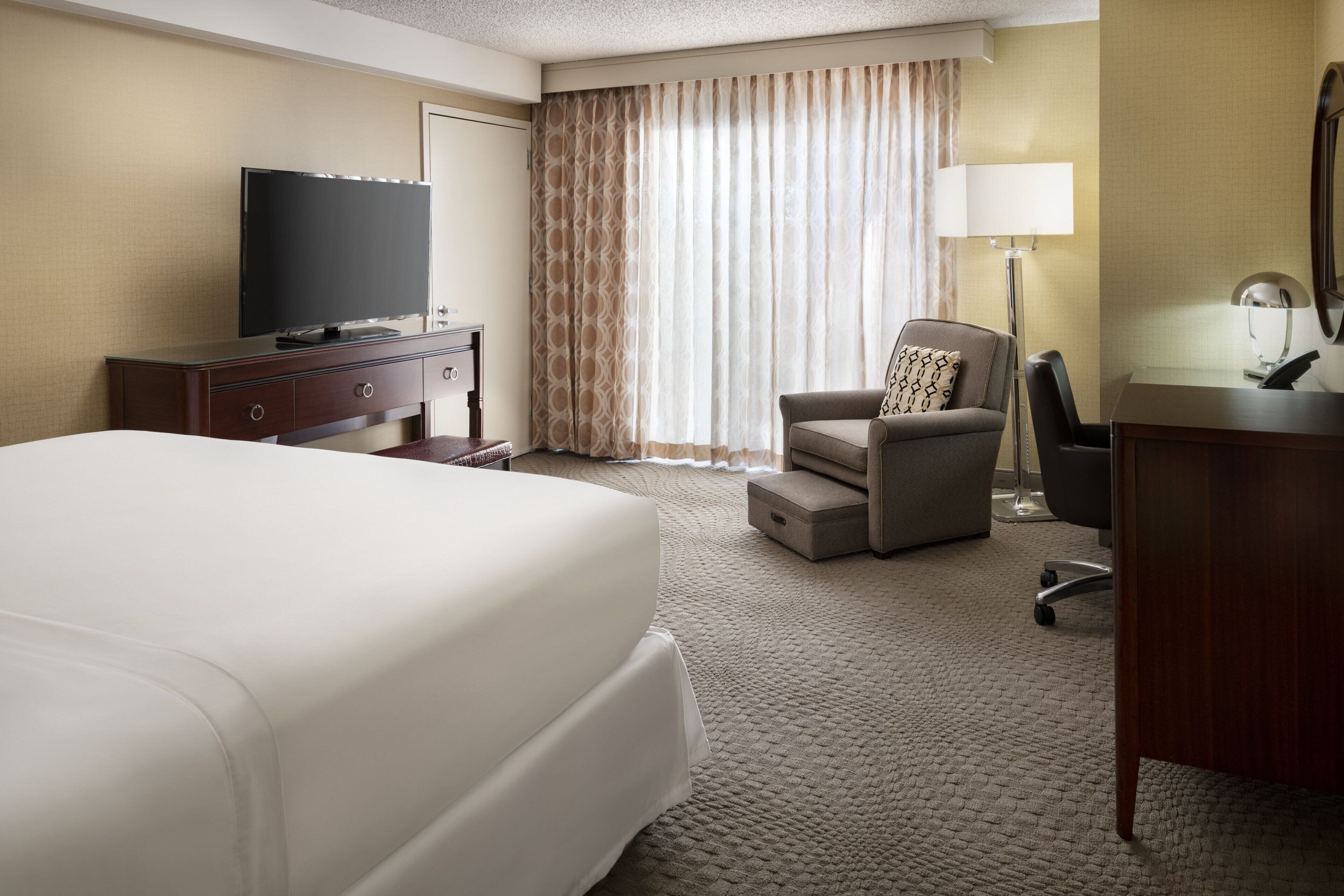 A hotel room with a large bed, a desk with a computer, a chair, a television, and a window with curtains.