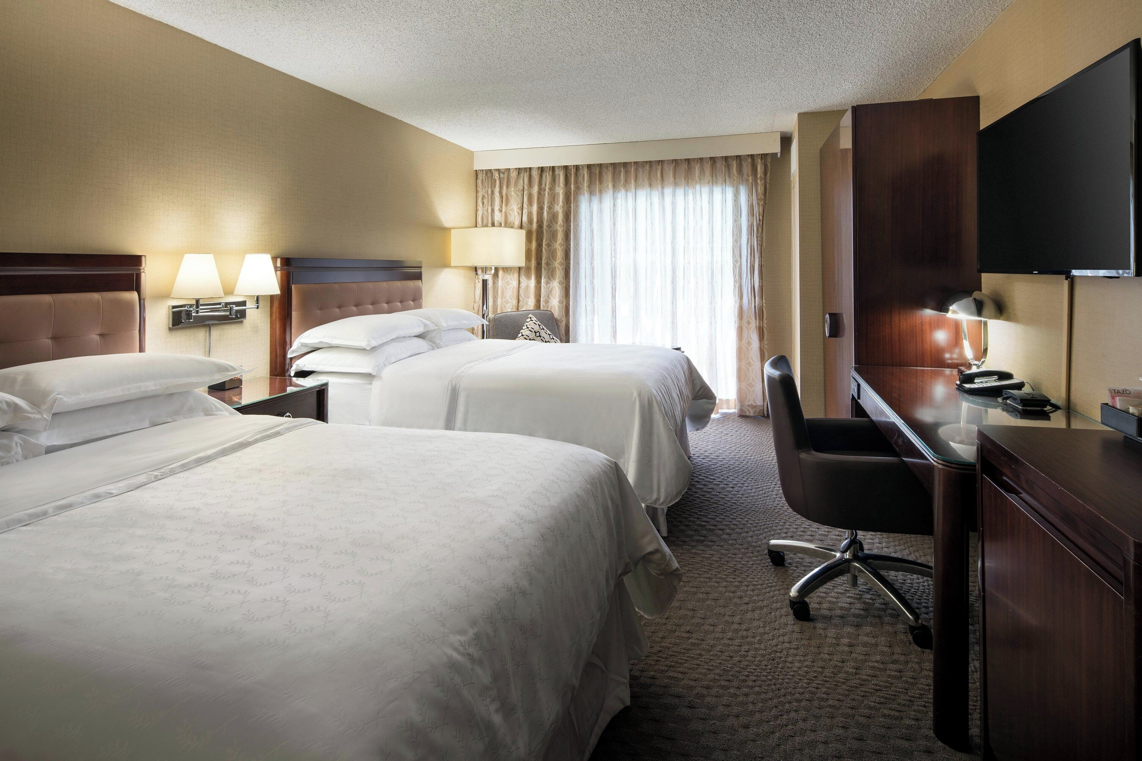 A hotel room with two beds, a desk, a chair, a TV, and a window with curtains.