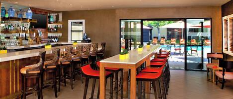 Zona de bar con taburetes de madera y una mesa larga, una barra con varias botellas y vistas a una piscina al aire libre.