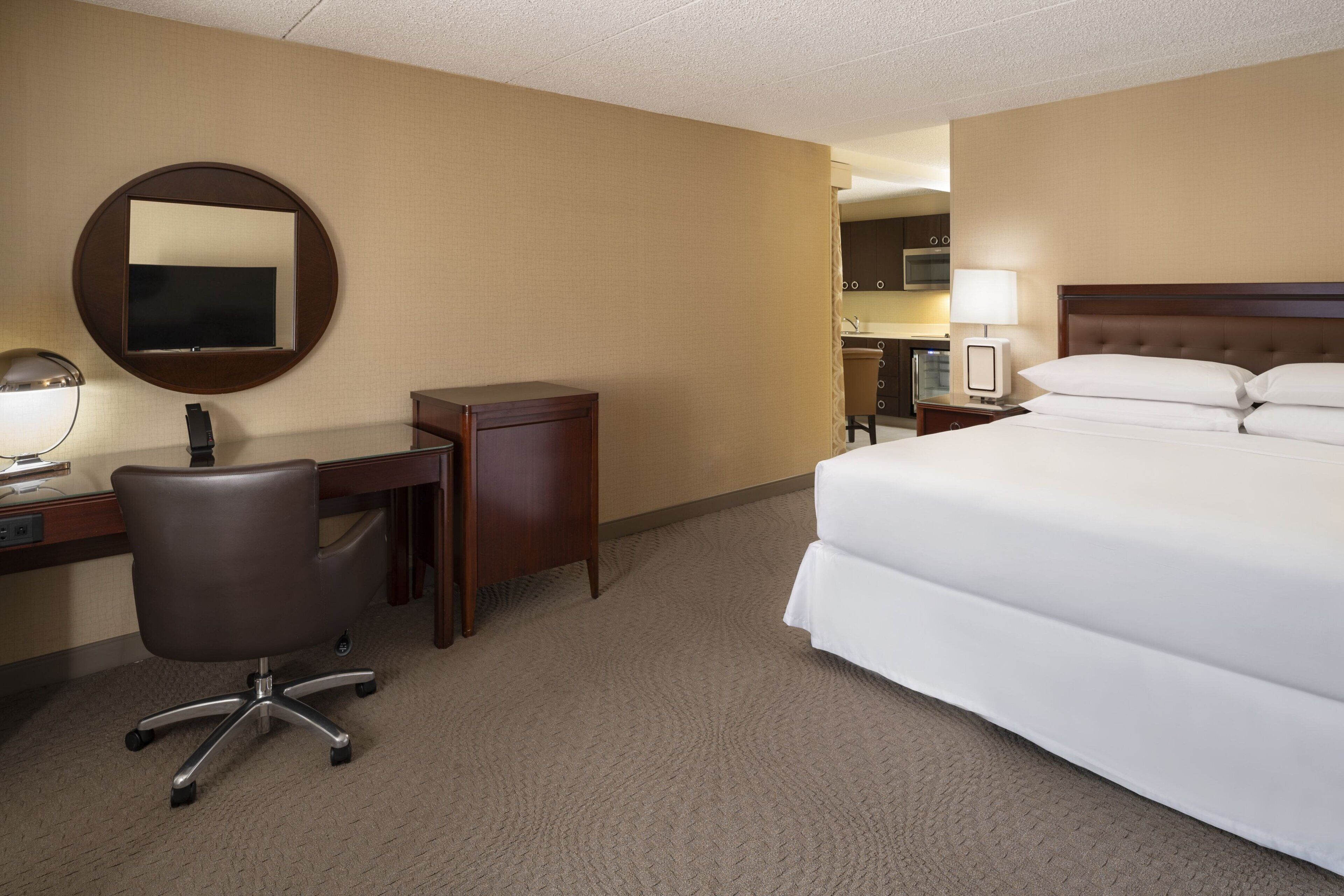 A hotel room with a bed, a desk with a chair, a round mirror, and a television.