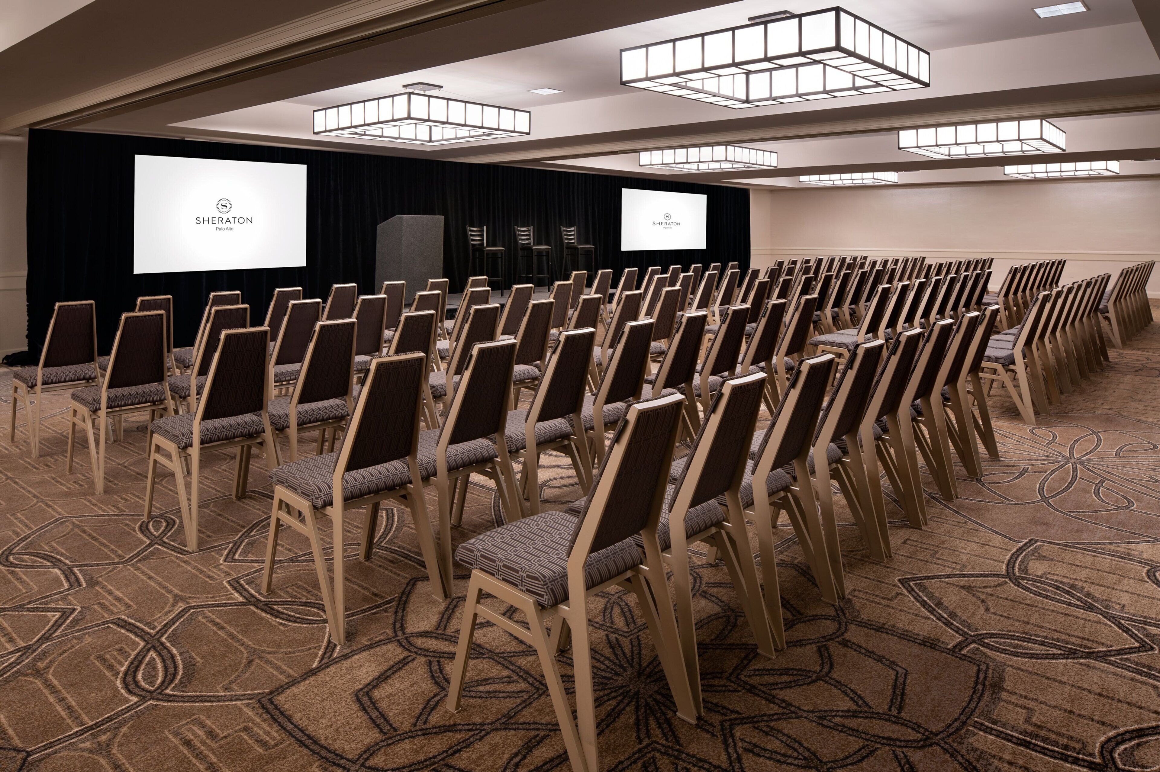 A conference room with rows of chairs, a stage with a projector screen, and a carpeted floor.