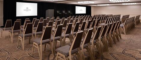A conference room with rows of chairs, a stage with a projector screen, and a carpeted floor.